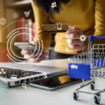 15 ventajas del comercio electrónico para las empresas | ¡Digitalízate! Una mujer con su celular y un computador junto a un pequeño carrito de compras y entre ellos íconos que muestran las ventajas del comercio electrónico